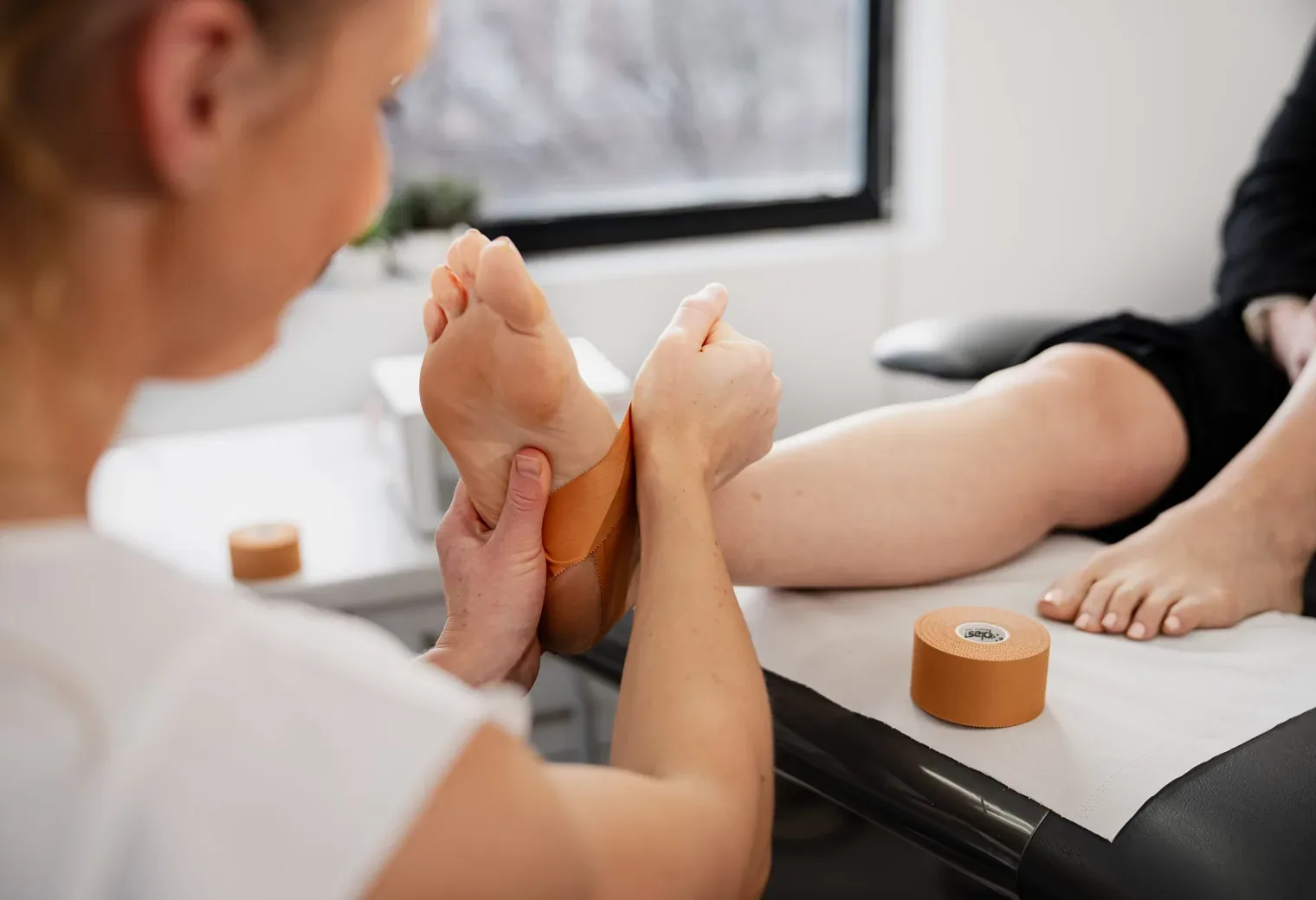 A podiatrist applying tan rigid strapping tape around the arch of a patient's bare foot, with a roll of tape on the treatment couch.