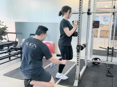 An Align Health Collective clinician kneeling beside a patient stepping up inside a squat rack.