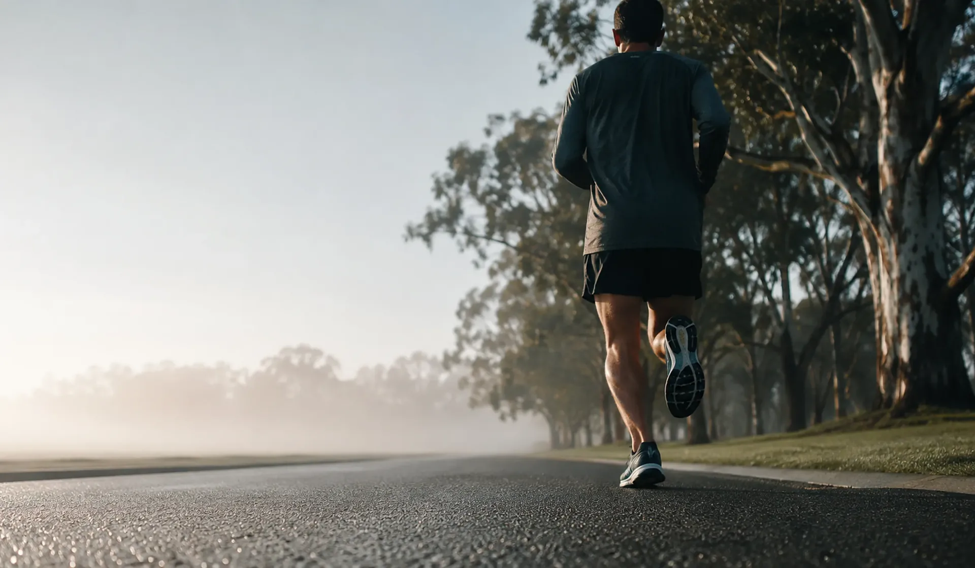 A runner seen from behind on an empty road at sunrise, mist over eucalypt trees along the roadside.