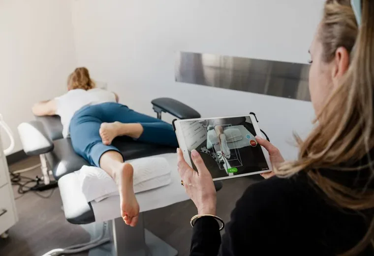 A treatment room with a patient lying face down on a couch, feet raised on a bolster, while a podiatrist scans their foot with a tablet.