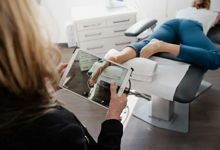 A podiatrist holding a tablet running a 3D foot-scanning app, with a patient lying on a treatment couch and their feet raised on a bolster.