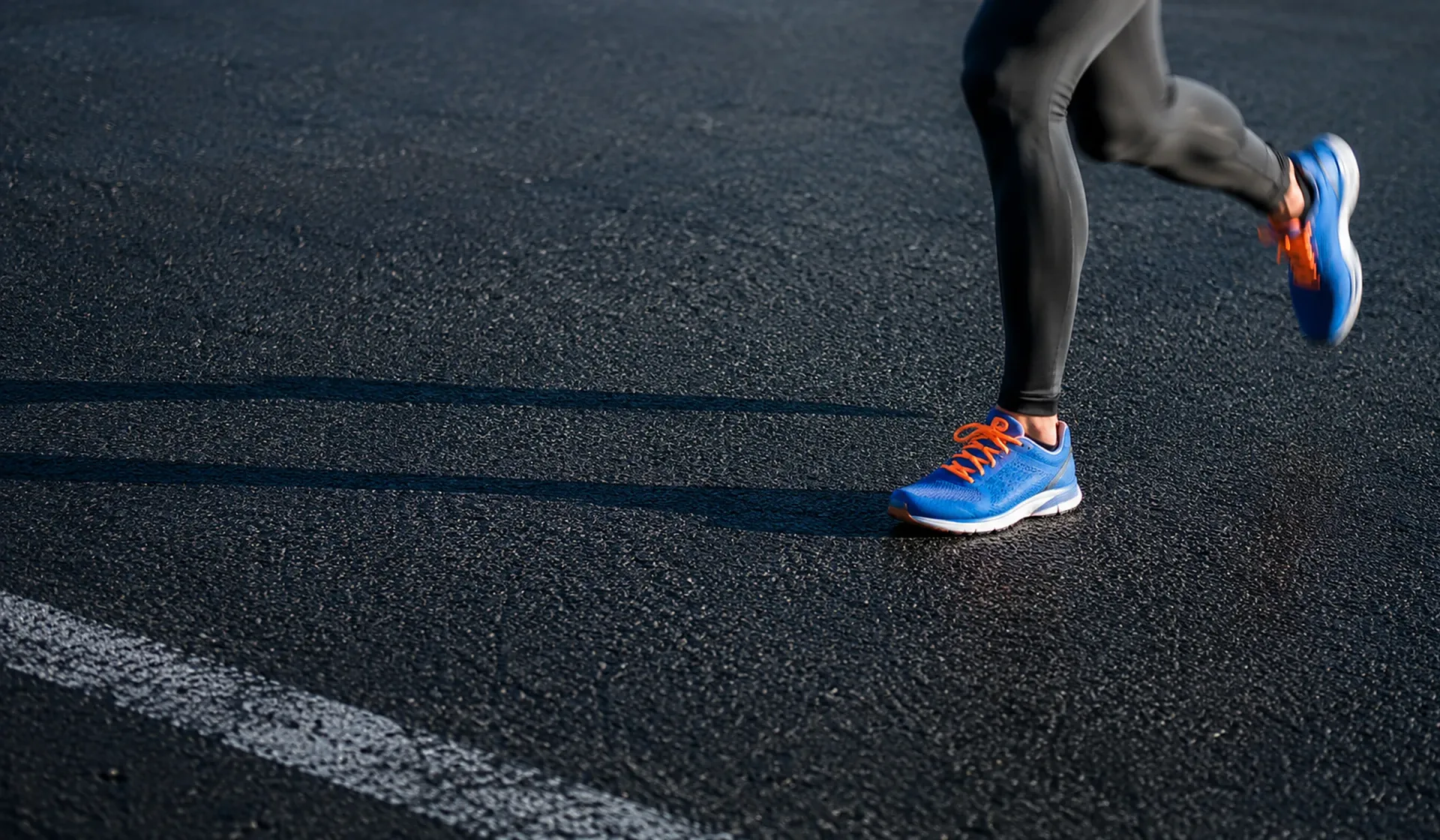 A runner mid-stride on dark asphalt at a low side angle, blue running shoes with orange laces and grey running tights, a white lane marking crossing the lower left of the road.